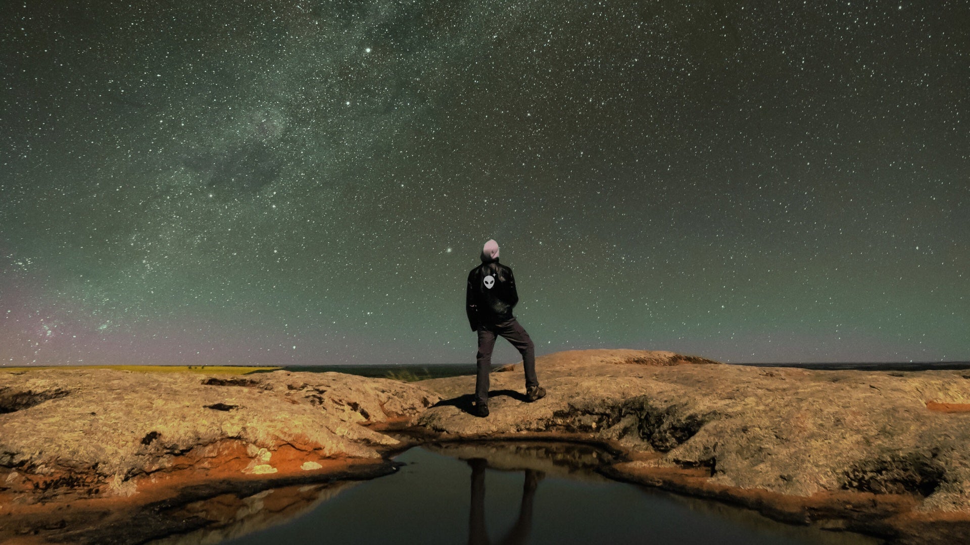 Person wearing a Stellarwear Alienhead jacket standing on a rocky outcrop under a starry night sky