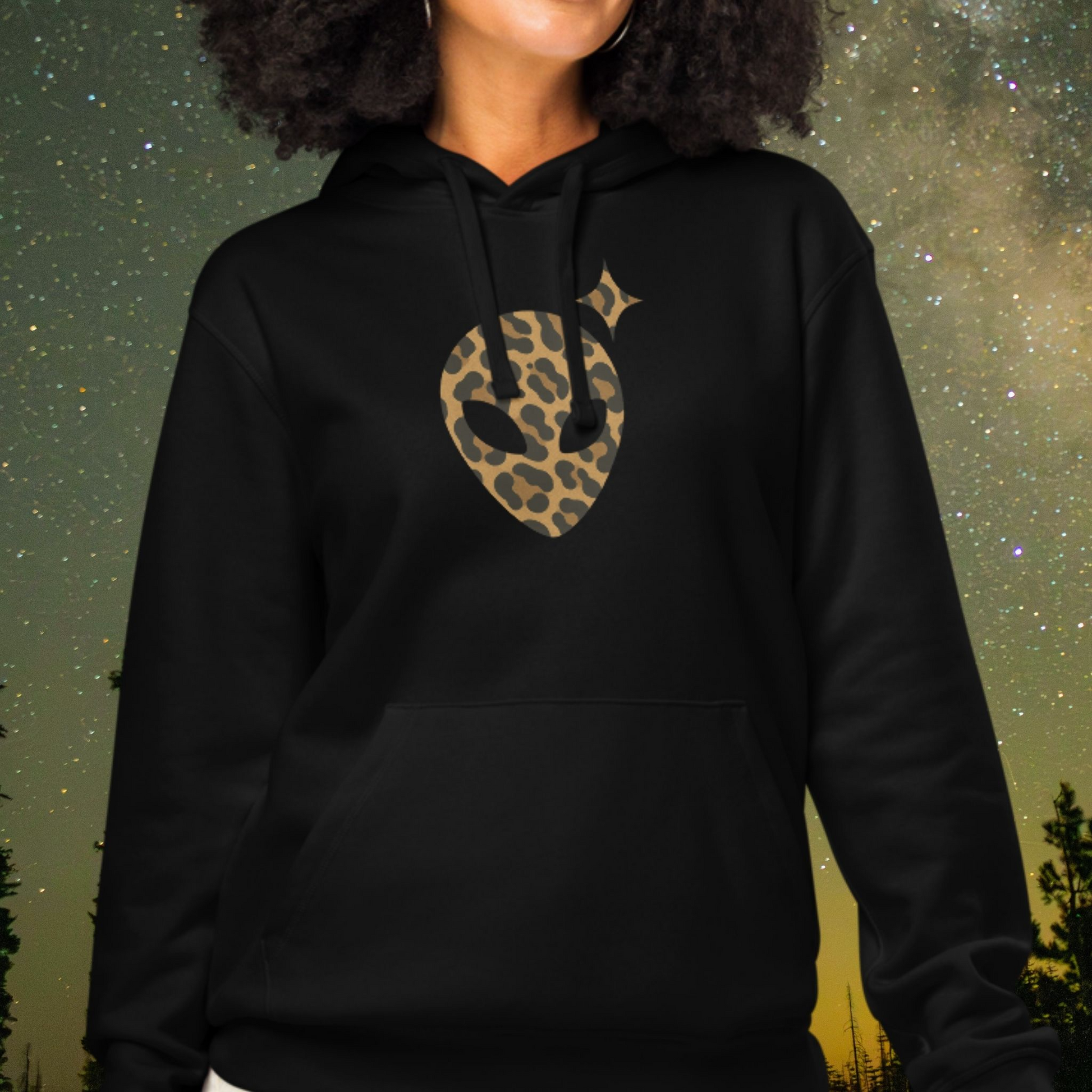 Person wearing a black Stellarwear hoodie with a leopard print alien head design against a starry night sky.
