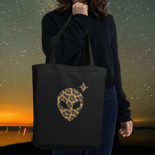 Person holding a black Stellarwear tote bag with an animal print Alienhead graphic against a night sky.