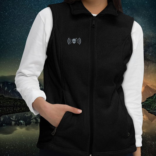 Person wearing a Stellarwear black fleece vest with an embroidered alien icon against a mountainous landscape background.