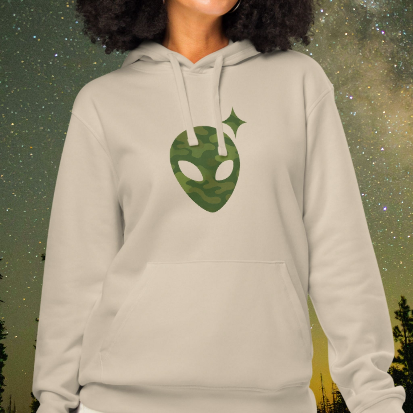 Person wearing a beige Stellarwear hoodie with a green alien head design against a starry night sky background