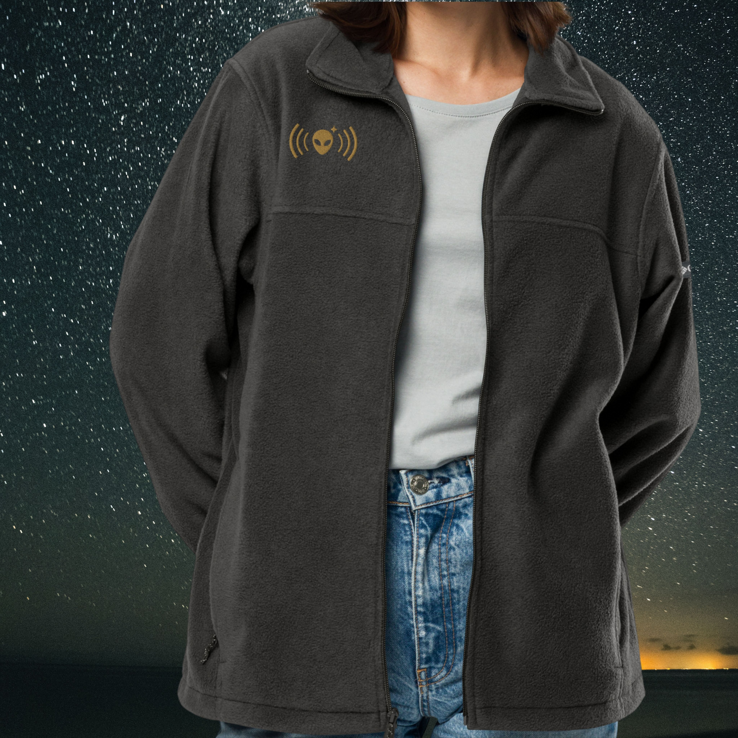 Person wearing a Stellarwear dark gray jacket with an alien signal icon on the chest against a starry night sky background.