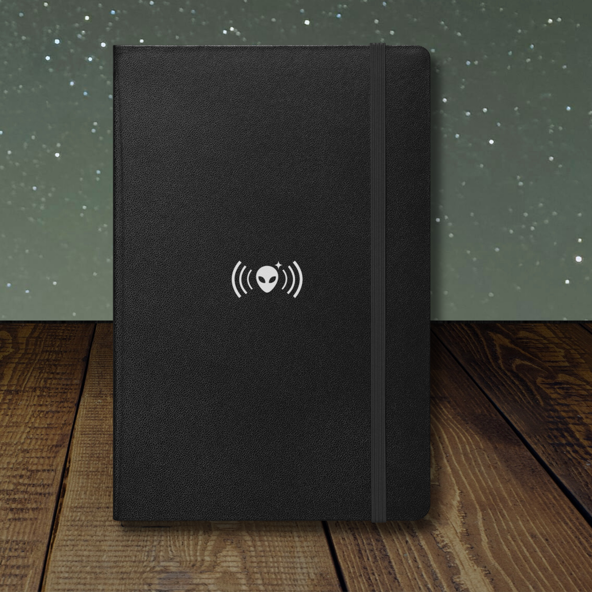 Stellarwear black notebook with a white alien logo on a wooden surface and starry background