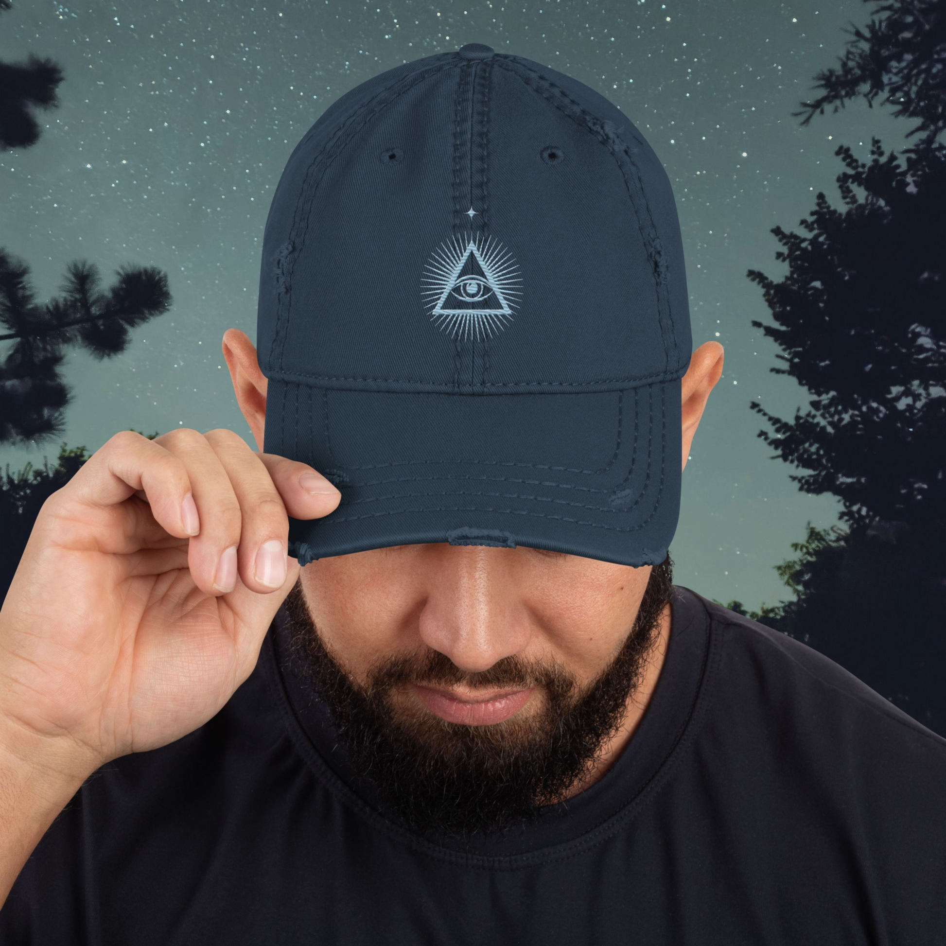 Man wearing a navy blue Stellarwear cap with a third eye logo against a night sky with trees