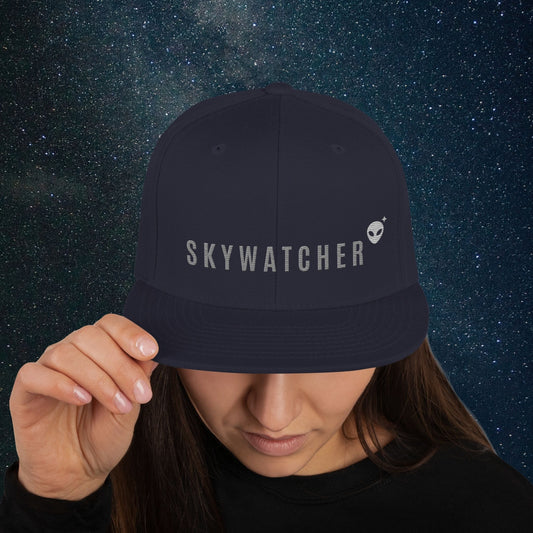 Person wearing a Stellarwear navy blue snapback cap with 'SKYWATCHER' logo against a starry night sky background